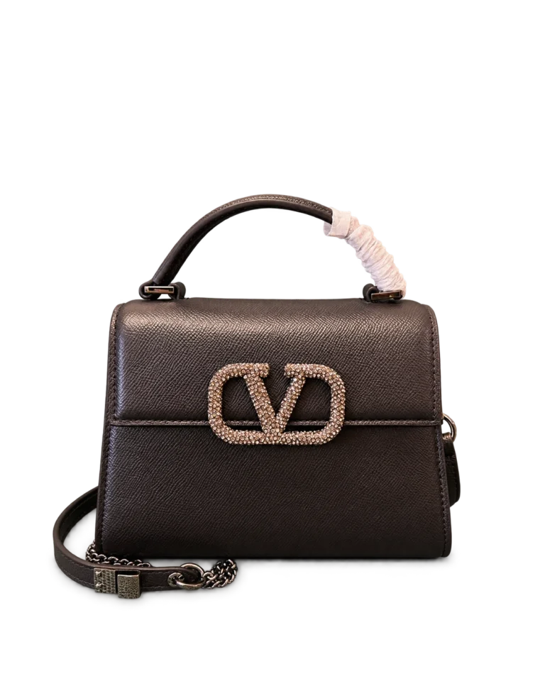Upright front view of a dark brown textured top-handle bag with a stone-covered emblem, shoulder strap and pink protective handle wrap
